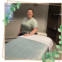 Ally Middleton, a Licensed Massage Therapist, sits smiling next to a professional massage table in her treatment room. The photo is bordered by a gold frame with green eucalyptus accents.