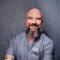Dr. Tom Ingegno smiling headshot in grey scrubs against a grey studio background.