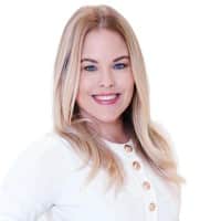 A professional studio headshot of Meghan Phelan, a blonde woman with blue eyes, smiling and wearing a white buttoned shirt against a clean white background.