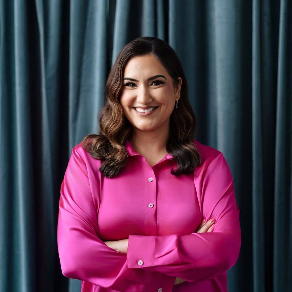 Professional headshot of Delisi Friday, founder of First Call Friday, smiling in a vibrant pink button-down shirt with her arms crossed against a dark draped curtain background.