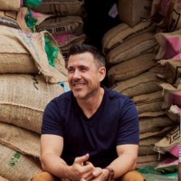 Paul Schlader, co-founder of Birch Coffee, sitting down and smiling in front of a large stack of burlap coffee bean sacks. He is wearing a dark blue v-neck shirt.