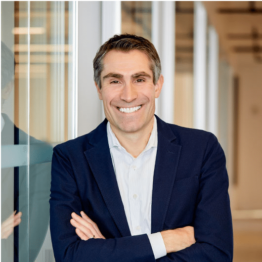 Corporate headshot of Michal Petrzela, Partner at Lightyear Capital, standing with arms crossed and smiling. He is wearing a dark navy blazer over an open-collar white dress shirt in a modern glass office hallway setting.